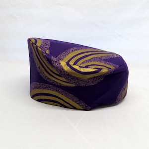 May include: A cylindrical pillbox hat in a deep purple fabric featuring a repeating pattern of curved, textured gold lines. The hat has a flat top and a structured, upright side profile, showcasing a smooth finish with a distinct metallic gold print.