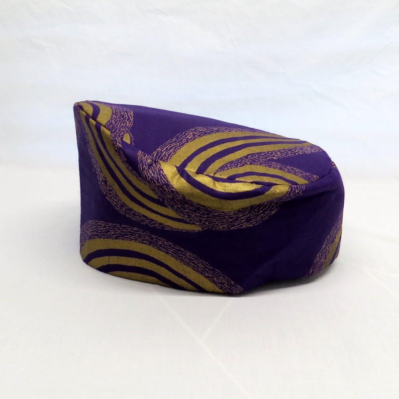 May include: A cylindrical pillbox hat in a deep purple fabric featuring a repeating pattern of curved, textured gold lines. The hat has a flat top and a structured, upright side profile, showcasing a smooth finish with a distinct metallic gold print.