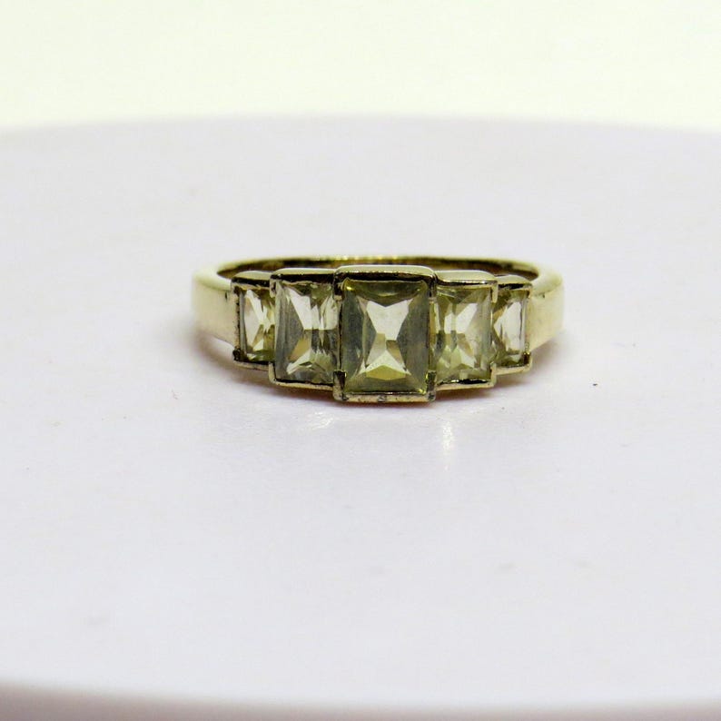 Op de afbeelding: Een goudkleurige ring met vijf rechthoekige, lichtgroene edelstenen. De edelstenen zijn in een rij over de bovenkant van de ring geplaatst. De ring heeft een eenvoudig, klassiek ontwerp, geschikt voor dagelijks gebruik of speciale gelegenheden.