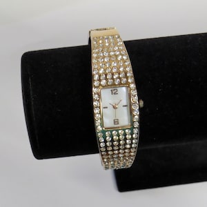 May include: A gold-toned bracelet watch with a rectangular face. The watch band and face are encrusted with clear rhinestones. The watch face has a white background with black numerals at the 12 and 6 positions and gold-toned hands.