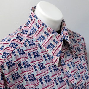 May include: A button-down shirt with a repeating pattern of figures in blue and red on a light blue and white background. The shirt is displayed on a white mannequin, showcasing the collar and a portion of the front.