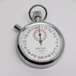 May include: A silver-toned analog stopwatch with a white face, featuring black and red numerals, a central sub-dial, and a winding crown with a top ring. The dial displays the text UNITED, 7 JEWELS, and 1/10, with SWISS MADE printed at the bottom.