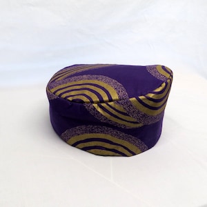 May include: A cylindrical hat in a deep purple fabric featuring a repeating pattern of concentric circles in a metallic gold finish. The hat has a flat top and a structured, upright side band, showcasing a consistent circular design throughout.
