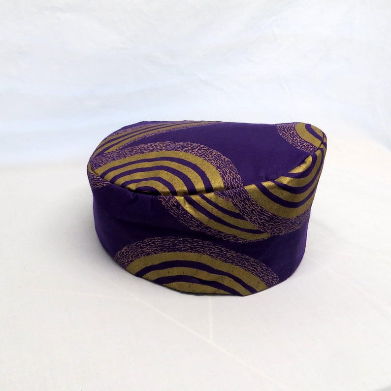 May include: A cylindrical hat in a deep purple fabric featuring a repeating pattern of concentric circles in a metallic gold finish. The hat has a flat top and a structured, upright side band, showcasing a consistent circular design throughout.