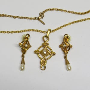 May include: Gold-toned jewelry set featuring a necklace with a pendant and matching earrings. The pendant and earrings have a Celtic knot design with a central pearl and a dangling pearl. The necklace chain is a delicate link style.