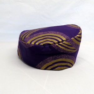 May include: A round, brimless hat in a deep purple fabric featuring a repeating pattern of concentric gold semi-circles. The hat has a structured, cylindrical shape with a flat top and a smooth, consistent finish across the entire exterior surface.