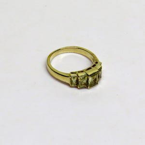 Op de afbeelding: Een goudkleurige ring met vijf rechthoekige, lichtgroene edelstenen. De ring heeft een eenvoudige band en de edelstenen zijn in een rij geplaatst. De ring wordt op een witte achtergrond getoond.