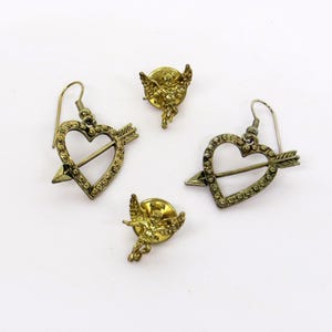 May include: A collection of vintage gold-toned jewelry, including two pairs of earrings with heart and arrow designs, and two small cherub pins. The earrings feature a heart pierced by an arrow, embellished with small stones.