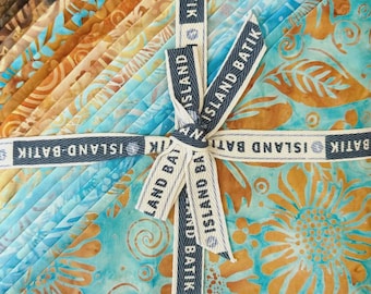 May include: A stack of folded fabric squares featuring batik patterns in shades of turquoise, teal, and golden brown. The fabric bundle is tied with a cream-colored ribbon printed with the text ISLAND BATIK in a repeating dark blue pattern.