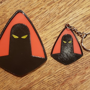 May include: Two 3D printed keychains featuring a black silhouette of a hooded figure with yellow eyes. The larger keychain is a standalone piece, while the smaller keychain is attached to a silver key ring.