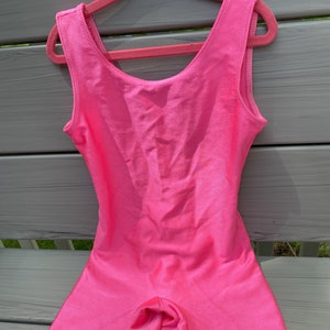 May include: A bright pink sleeveless leotard with a short leg design. The leotard is made of a shiny, smooth fabric.