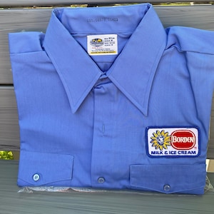 May include: A light blue work shirt with a white and blue Borden Milk & Ice Cream logo patch. The shirt has a button-down collar, two chest pockets, and a label that reads "LOT. 00679 10423".