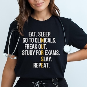 May include: Black t-shirt with white and yellow text that reads "EAT. SLEEP. GO TO CLINICALS. FREAK OUT. STUDY FOR EXAMS. SLAY. REPEAT." A stethoscope hangs around the neck. The shirt is paired with black pants.