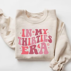 May include: A beige sweatshirt with the text "IN MY THIRTIES ERA" in pink and red retro font with a butterfly and stars.