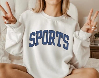 May include: White sweatshirt with a navy blue "SPORTS" graphic on the front. The sweatshirt is worn by a person sitting on a bed.