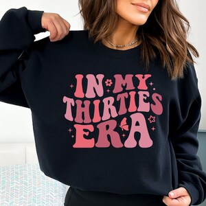 May include: Black sweatshirt with a pink and red graphic that says "In My Thirties Era" in a retro font.