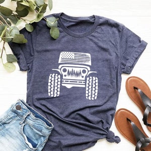 May include: A navy blue t-shirt with a white graphic of a Jeep with an American flag on the hood. The Jeep is driving through a forest.