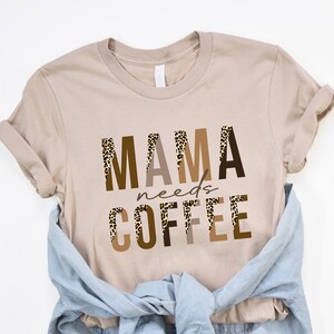 Op de afbeelding: T-shirt in tan met een grafische print die "MAMA needs COFFEE" zegt in bruine en zwarte letters met een luipaardprint.