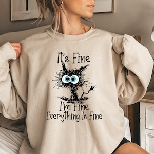 May include: A beige sweatshirt with a black cat illustration and the text "It's Fine I'm Fine Everything Is Fine".