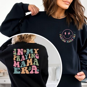 May include: Black sweatshirt with a graphic on the front that says "Praying Mama Era" with a smiley face and lightning bolt. The back of the sweatshirt has a graphic that says "In My Praying Mama Era" with flowers and a butterfly.