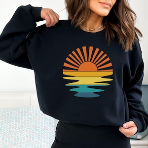 May include: Black sweatshirt featuring a retro sunset design. The graphic includes a sun with orange and yellow rays, reflected in layered bands of yellow, teal, and blue. A casual, comfortable top for everyday wear.