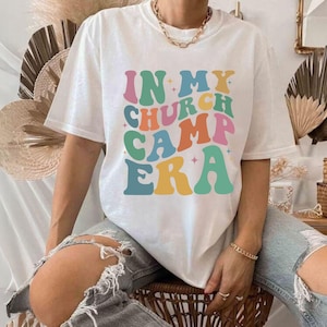 K&ouml;nnte beinhalten: Wei&szlig;es T-Shirt mit einer Retro-Grafik, auf der "In My Church Camp Era" in bunten Buchstaben steht.