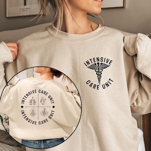 May include: Beige sweatshirt with black text and graphics. The front features the words "INTENSIVE CARE UNIT" with a caduceus symbol. The back has a circular design with the same text and medical illustrations. A comfortable, casual garment.