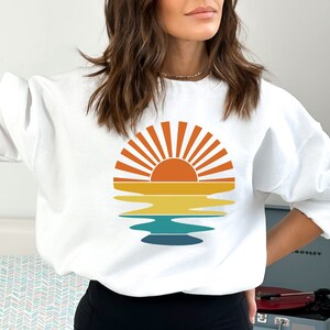 May include: White sweatshirt featuring a retro sunset design. The graphic includes a sun with orange and yellow rays, a yellow and orange horizon line, and a blue and teal water reflection. A casual, comfortable top for everyday wear.