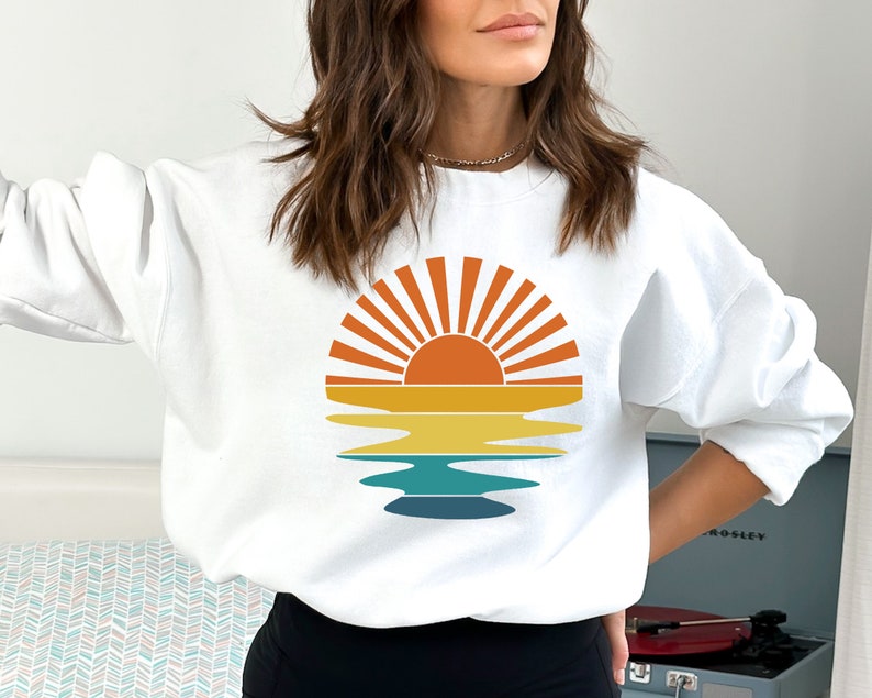 May include: White sweatshirt featuring a retro sunset design. The graphic includes a sun with orange and yellow rays, a yellow and orange horizon line, and a blue and teal water reflection. A casual, comfortable top for everyday wear.