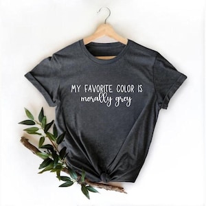 May include: A dark charcoal grey short-sleeved t-shirt hanging on a wooden hanger. The shirt features white printed text that reads, "MY FAVORITE COLOR IS morally grey" in a combination of block and script fonts centered on the chest area.