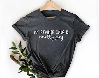 May include: A dark charcoal grey short-sleeved t-shirt hanging on a wooden hanger. The shirt features white printed text that reads, "MY FAVORITE COLOR IS morally grey" in a combination of block and script fonts centered on the chest area.