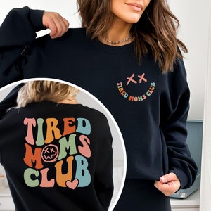 Puede incluir: Sudadera negra con el texto "Tired Moms Club" en colores arco&iacute;ris y una carita sonriente con una X por ojos en la parte delantera. La parte posterior de la sudadera tiene el mismo texto en letras arco&iacute;ris m&aacute;s grandes con una carita sonriente con una X por ojos.