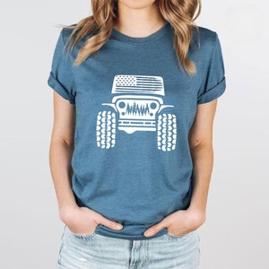 May include: A blue t-shirt with a white graphic of a Jeep with an American flag on the hood and trees in the grill. The Jeep has large tires.