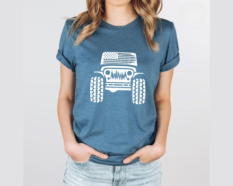 May include: A blue t-shirt with a white graphic of a Jeep with an American flag on the hood and trees in the grill. The Jeep has large tires.