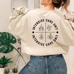May include: Cream-colored sweatshirt with a circular graphic on the back. The graphic features anatomical illustrations of the lungs, heart, brain, and kidneys, with the words "INTENSIVE CARE UNIT" encircling the images.