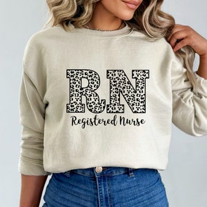 May include: A beige sweatshirt with a black and white leopard print "RN" design and the text "Registered Nurse" printed on the front.