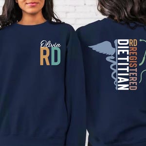 May include: Navy blue sweatshirt with "Olivia RD" in white script and orange and teal block letters. The back features a caduceus symbol, a stethoscope, and the words "Registered Dietitian" in white and coral.