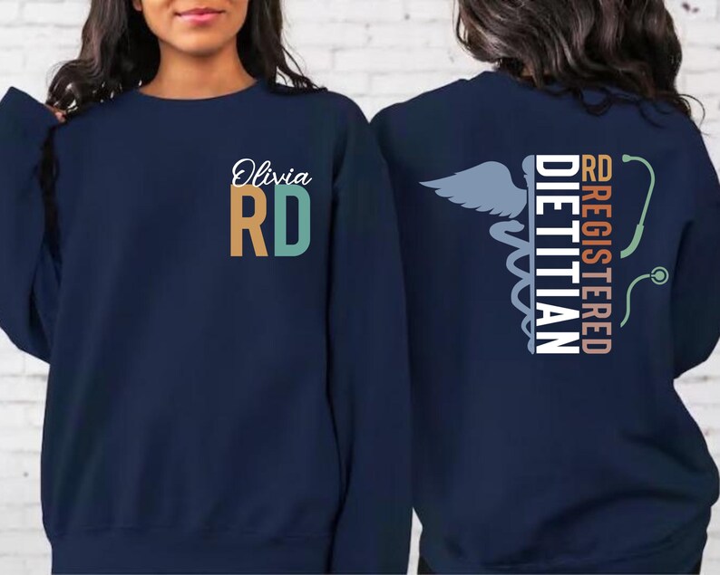 May include: Navy blue sweatshirt with "Olivia RD" in white script and orange and teal block letters. The back features a caduceus symbol, a stethoscope, and the words "Registered Dietitian" in white and coral.