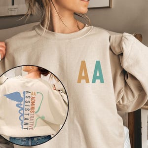 May include: A beige sweatshirt with the letters "AA" in gold and teal on the front. The back of the sweatshirt features a medical caduceus with the words "Administrative Assistant" in teal and gold.