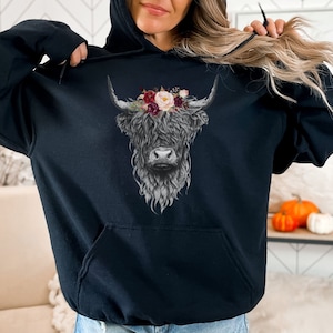 Puede incluir: Sudadera negra con un gr&aacute;fico de una vaca de las tierras altas con una corona de flores. La vaca est&aacute; de frente y tiene la cara blanca y los cuernos negros.