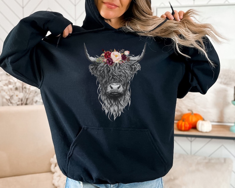 Puede incluir: Sudadera negra con un gr&aacute;fico de una vaca de las tierras altas con una corona de flores. La vaca est&aacute; de frente y tiene la cara blanca y los cuernos negros.