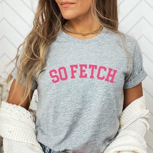 Op de afbeelding: Grijs T-shirt met de tekst "SO FETCH" in roze letters.
