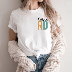 Puede incluir: Camiseta blanca con el texto "Olivia RD" en negro, dorado y turquesa. La camiseta est&aacute; anudada a la cintura y combinada con vaqueros azules. Un su&eacute;ter color crema est&aacute; drapeado sobre los hombros.