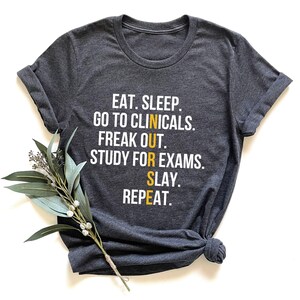 May include: Dark gray t-shirt with rolled sleeves and a knotted hem. The shirt features white and yellow text that reads: "EAT. SLEEP. GO TO CLINICALS. FREAK OUT. STUDY FOR EXAMS. SLAY. REPEAT." A sprig of greenery rests on the shirt.