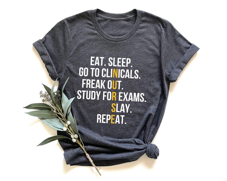 May include: Dark gray t-shirt with rolled sleeves and a knotted hem. The shirt features white and yellow text that reads: "EAT. SLEEP. GO TO CLINICALS. FREAK OUT. STUDY FOR EXAMS. SLAY. REPEAT." A sprig of greenery rests on the shirt.