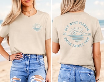 May include: A beige short-sleeve t-shirt featuring a small blue sun graphic on the front left chest. The back displays a large circular blue graphic with a sun, water, and the text "KEY WEST FLORIDA 2026 SMITH FAMILY TRIP" in a circular layout.
