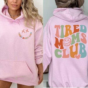 Puede incluir: Sudadera rosa con capucha con el texto "TIRED MOMS CLUB" en colores arco&iacute;ris en la parte posterior y una carita sonriente con una X por ojos y el texto "TIRED MOMS CLUB" en colores arco&iacute;ris en la parte delantera.