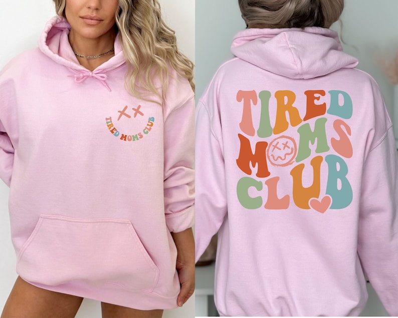 Puede incluir: Sudadera rosa con capucha con el texto "TIRED MOMS CLUB" en colores arco&iacute;ris en la parte posterior y una carita sonriente con una X por ojos y el texto "TIRED MOMS CLUB" en colores arco&iacute;ris en la parte delantera.