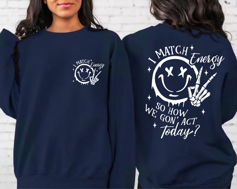 May include: A navy blue sweatshirt with a white graphic of a smiley face with crossed eyes and a skeleton hand giving the peace sign. The text "I Match Energy" is above the graphic and the text "So How We Gon' Act Today?" is below the graphic.