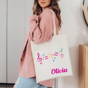 Może przedstawiać: Biała torba z pł&oacute;tna z kolorowymi nutami i kluczem wiolinowym. Torba jest spersonalizowana z imieniem "Olivia" nadrukowanym na r&oacute;żowo.